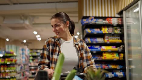 A-happy-brunette-girl-in-a-plaid-shirt-and-a-white-T-shirt-walks-along-the-shelves-and-inspects-goods-in-a-supermarket