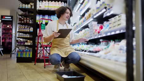 A-guy-with-curly-hair-with-a-tablet-in-his-hands-takes-inventory-of-goods-in-the-dairy-products-compartment-in-a-supermarket