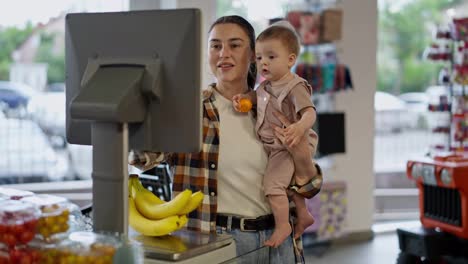 Glückliches-Brünettes-Mädchen,-Das-Ihr-Kleines-Baby-In-Den-Armen-Hält-Und-Bananen-Mit-Einer-Waage-In-Einem-Supermarkt-Wiegt