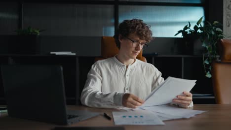 A-confident-young-guy-with-curly-hair-in-glasses-and-a-white-shirt-goes-through-documents-and-examines-papers-while-sitting-at-a-table-in-the-office