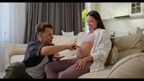 A-happy-man-and-his-pregnant-wife-are-having-fun-and-painting-the-sun-on-their-stomach-with-yellow-paint-at-home-on-the-sofa