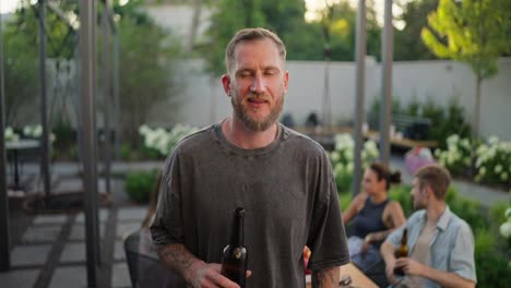 Portrait-of-a-confident-blond-guy-with-a-beard-in-a-gray-T-shirt-holding-a-brown-bottle-of-beer-and-posing-in-the-yard-of-a-back-house-near-his-company-on-vacation