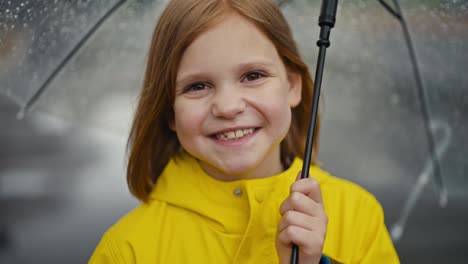 Portrait-of-a-happy-blonde-teenage-girl-in-a-yellow-jacket-holding-a-transparent-umbrella-with-water-drops-in-the-park-while-walking-after-the-rain