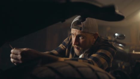 Close-up-a-mechanic-guy-in-a-checkered-shirt-and-cap-repairs-a-motorcycle-with-a-screwdriver-in-an-repair-shop