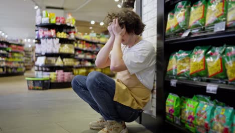 Un-Chico-Cansado-Con-El-Pelo-Rizado,-Trabajador-De-Un-Supermercado,-Se-Apoya-En-La-Pared-Cerca-De-Los-Estantes-Con-Productos-Y-Se-Toma-Un-Descanso-Durante-Un-Día-Ajetreado-En-El-Trabajo.