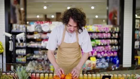 Confident-brunette-supermarket-worker-with-curly-hair-in-an-apron-lays-out-citrus-and-orange-fruits-on-a-shelf-in-a-supermarket