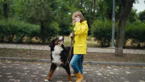 Side-view-of-a-happy-little-blonde-girl-in-a-yellow-jacket-walking-with-her-dog-along-the-alley-in-the-park-after-the-rain