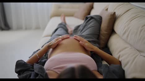 Top-view-of-happy-pregnant-brunette-woman-stroking-her-belly-while-lying-on-sofa-at-home