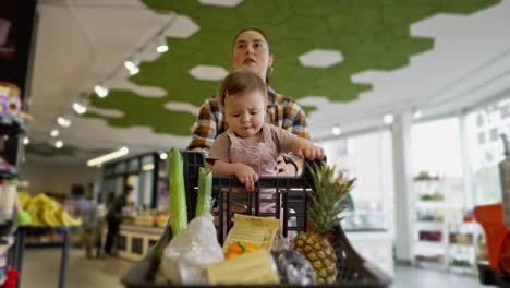 A-little-baby-girl-sits-in-a-cart-and-touches-with-her-hands-the-goods-that-she-and-her-mother-chose-while-shopping-in-a-supermarket