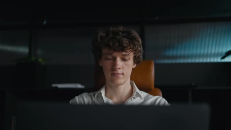 Close-up-of-a-confident-guy-with-curly-hair-in-a-white-shirt-working-using-a-gray-laptop-while-sitting-at-a-table-in-the-office