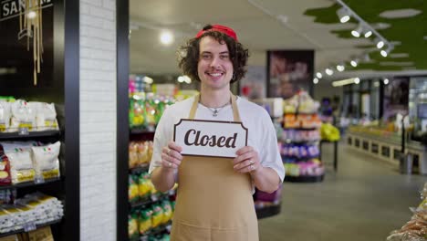 A-smiling-guy-with-curly-hair-and-yellow-front-stands-in-a-supermarket-and-holds-a-sign-in-front-of-his-chest-that-says-Closed