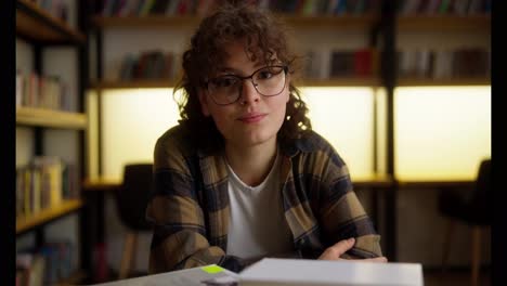 Contemplation-of-a-happy-girl-student-with-curly-hair-and-piercing-who-sits-at-a-table-in-the-library-among-books