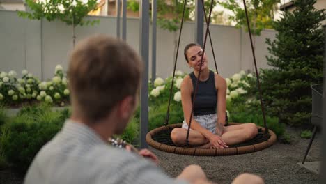 Over-the-shoulder-a-happy-brunette-girl-swings-on-a-rope-swing-and-sings-a-song-together-with-her-boyfriend-who-plays-the-guitar-in-the-backyard-of-a-country-house