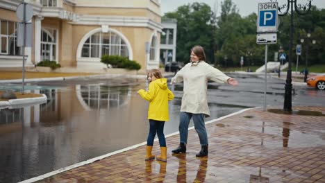 A-happy-woman-in-a-white-jacket-together-with-her-teenage-daughter-in-a-yellow-jacket-are-standing-on-the-edge-of-the-road-and-they-are-doused-with-water-by-a-car-driving-through-a-puddle-after-the-rain