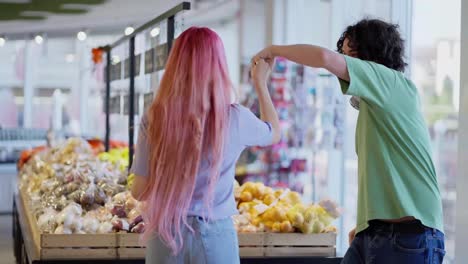 A-happy-girl-with-pink-hair-dances-with-a-brunette-guy-in-a-green-T-shirt-near-the-counter-in-a-supermarket