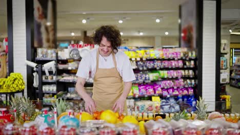 Glücklicher-Kerl-Mit-Lockigem-Brünettem-Haar-Supermarktangestellter-Legt-Obst-Ins-Regal-Während-Seiner-Arbeit-Im-Supermarkt