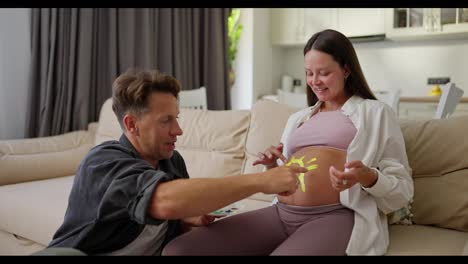 Happy-brunette-pregnant-woman-paints-a-sun-on-her-belly-with-yellow-matching-paint-together-with-her-husband-while-expecting-a-baby-at-home