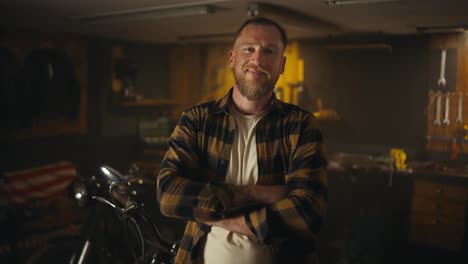 Portrait-of-a-happy-blond-guy-with-a-beard-in-a-checkered-shirt-who-crossed-his-arms-over-his-chest-and-poses-in-his-workshop