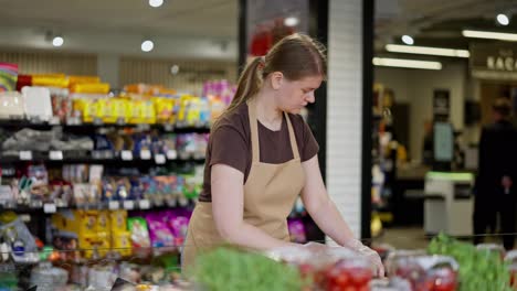 Confident-girl-in-a-brown-T-shirt-and-apron-lays-out-vegetables-and-fruits-on-the-counter-in-a-supermarket-during-her-working-day