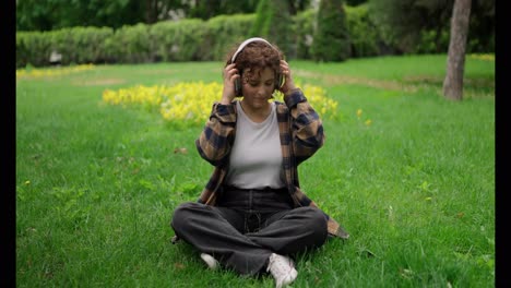 Happy-brunette-girl-in-a-brown-shirt-sits-on-the-lawn-and-turns-on-music-on-wireless-headphones-in-the-park