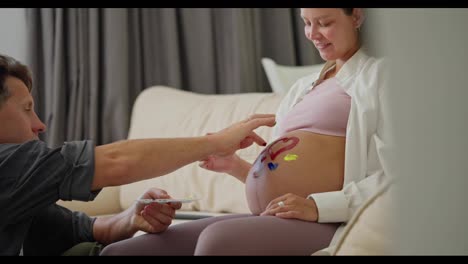 Side-view-of-a-happy-man-and-his-pregnant-wife-painting-on-their-belly-with-bright-colors-while-dreaming-about-a-future-child-and-expecting-a-child-at-home