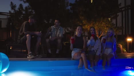 Un-Grupo-De-Amigos-Alegres-Chocando-Sus-Copas,-Charlando-Y-Bebiendo-Cerca-De-La-Piscina-Por-La-Noche.