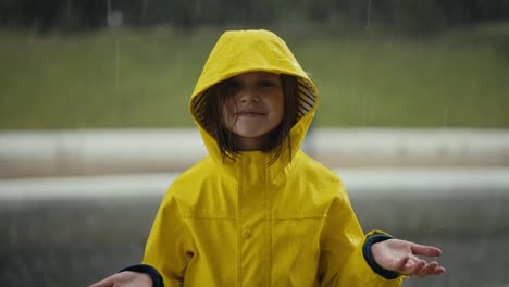 Portrait-of-a-happy-little-teenage-girl-in-a-yellow-jacket-standing-in-the-rain-and-catching-raindrops-with-her-hands-in-the-park-while-walking