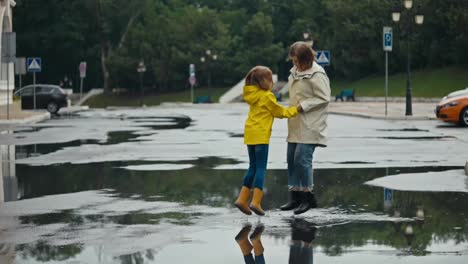 Mujer-Rubia-Feliz-Con-Su-Hija-Adolescente-Con-Una-Chaqueta-Amarilla-Saltando-En-Un-Charco-Tomadas-De-La-Mano-En-El-Parque-Después-De-La-Lluvia