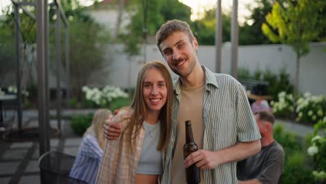 Portrait-of-a-happy-blond-guy-with-his-girlfriend-in-a-blue-top-who-is-hugging-the-guy-and-posing-in-the-courtyard-of-a-country-house