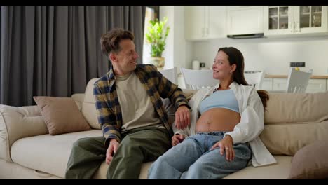 Happy-middle-aged-man-in-a-plaid-shirt-sits-on-the-sofa-with-his-pregnant-wife-and-talks-about-the-unborn-child-at-home