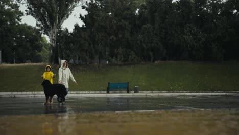 Side-view-of-a-happy-teenage-girl-in-a-yellow-jacket-playing-with-her-mom-and-a-black-dog-during-the-rain-in-the-park