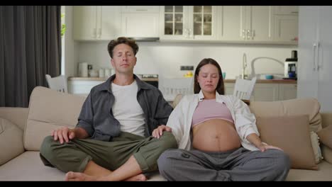 Confident-middle-aged-man-along-with-his-pregnant-wife-sit-on-the-sofa-and-meditate-during-their-leisure-time-at-home-on-the-sofa-while-waiting-for-the-baby-at-home