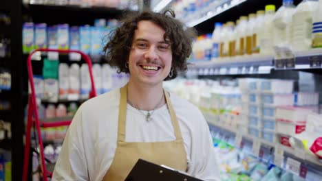 Portrait-of-a-happy-guy-with-curly-hair-in-a-yellow-apron-doing-inventory-and-holding-a-tablet-in-his-hands-in-the-dairy-section-of-a-supermarket