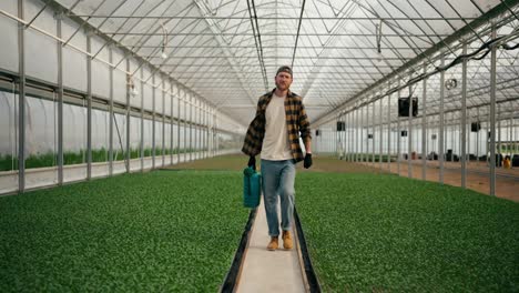 Portrait-of-a-confident-guy-in-a-farmer-cap-walking-along-plant-sprouts-and-carrying-a-blue-watering-can-in-his-hands-while-working-in-a-greenhouse-on-a-farm