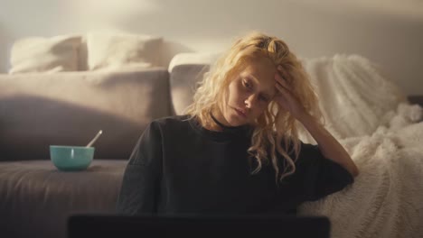 A-tired-blonde-girl-in-a-gray-T-shirt-leans-on-her-hand-and-looks-sadly-at-the-laptop-screen-near-the-sofa-at-home