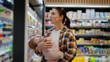 A-brunette-girl-in-a-plaid-shirt-holds-her-infant-child-in-her-arms-and-looks-at-the-goods-on-the-counter-in-the-dairy-department-of-a-supermarket