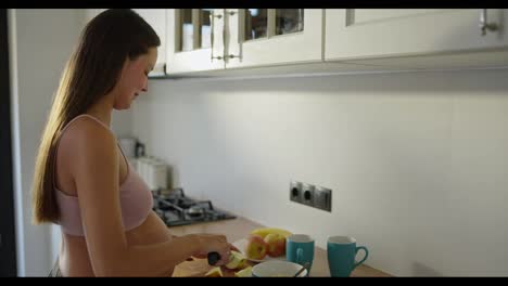 Happy-pregnant-brunette-woman-cutting-apples-for-fruit-salad-in-the-morning-during-breakfast-in-the-kitchen