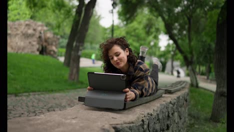 Happy-girl-in-a-plaid-shirt-lies-on-a-bench-and-moves-her-legs-while-watching-lessons-on-a-tablet-in-the-park