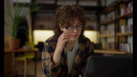 A-tired-girl-student-with-curly-hair-takes-off-her-glasses-during-her-studies-at-the-laptop-at-the-table-in-the-library