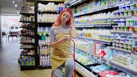 Ein-Müdes-Mädchen-Mit-Rosa-Haaren-Macht-Eine-Pause-Und-Scrollt-Auf-Ihrem-Telefon-Durch-Soziale-Netzwerke,-Während-Sie-In-Der-Milchprodukteabteilung-Eines-Supermarkts-Arbeitet