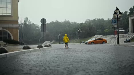 Rückansicht-Eines-Glücklichen-Teenager-Mädchens-In-Einer-Gelben-Jacke,-Das-Bei-Starkem-Regen-Durch-Pfützen-Und-Wasser-Im-Park-Läuft