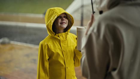 Über-Der-Schulter-Hält-Ein-Glückliches-Junges-Mädchen-In-Einer-Gelben-Jacke-Eine-Flasche-Wasser-Und-Kommuniziert-Mit-Ihrer-Mutter,-Während-Sie-Im-Regen-Im-Park-Spazieren-Geht