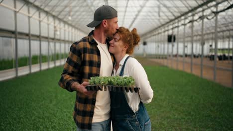 Una-Pareja-De-Agricultores-Enamorados,-Un-Chico-Rubio-Y-Su-Esposa-Pelirroja,-Posan-En-Un-Invernadero-Entre-Los-Brotes-De-Plantas-Jóvenes-En-La-Granja.