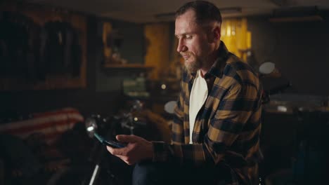 Confident-blond-guy-in-a-checkered-shirt-scrolls-social-networks-on-his-smartphone-in-a-workshop-garage