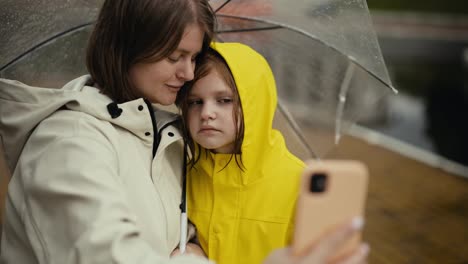 Happy-blonde-woman-in-a-white-jacket-takes-a-selfie-with-her-teenage-daughter-in-a-yellow-jacket-and-holds-an-umbrella-while-walking-after-the-rain-in-the-park