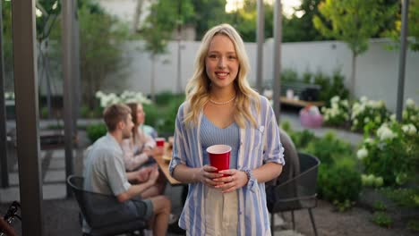 Portrait-of-a-happy-blonde-girl-in-a-blue-shirt-with-a-red-plastic-glass-in-her-hands-who-poses-and-smiles-while-relaxing-with-her-friends-in-the-courtyard-of-the-house