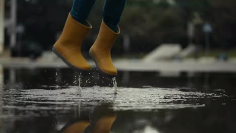 Close-up-of-a-teenage-girl-in-orange-rubber-boots-jumping-in-a-puddle-and-splashing-water-after-rain-in-the-park