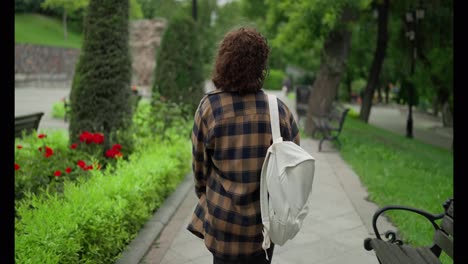 Back-view-A-brunette-girl-in-a-checkered-shirt-with-a-white-backpack-walks-through-the-park-after-classes-at-the-university