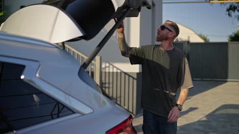 Confident-guy-in-sunglasses-with-his-girlfriend-putting-bags-in-the-trunk-of-the-car-and-getting-into-the-cabin-while-leaving-the-hotel-in-summer