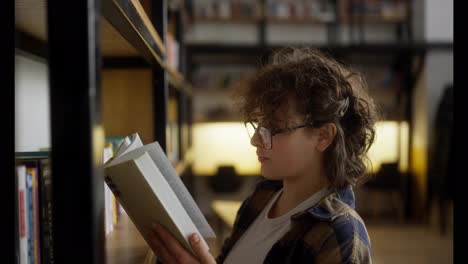 Confident-girl-with-curly-hair-with-glasses-in-a-checkered-shirt-reads-a-book-near-a-shelf-with-books-in-a-university-library